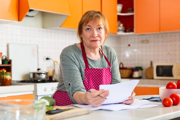 Fototapeta na wymiar Worried elderly housewife sitting in home kitchen, checking utility bills.