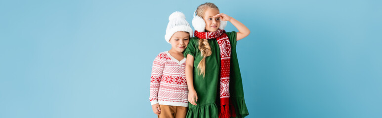 panoramic shot of girl in winter earmuffs looking away while standing near brother in hat on blue