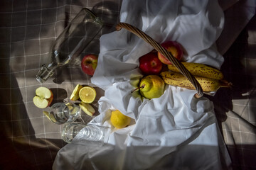 ripe fruits red apples, green pear, lemons, bananas in a basket with white drapery with folds, on checkered blanket with glass and a bottle and apple slices in sunlight
