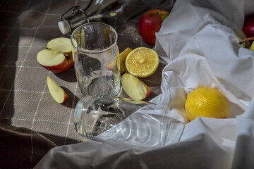 ripe fruits red apples, cut lemons on white drapery with folds, on checkered blanket with glass and a bottle and apple slices in sunlight