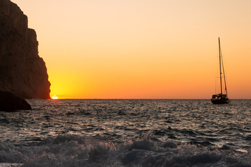 Sunset over the sea horizon with a sailboat next to a cliff in Majorca. Vacations concept