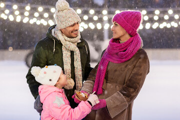 christmas, family and leisure concept - happy mother, father and daughter eating takeaway pancakes at outdoor skating rink in winter