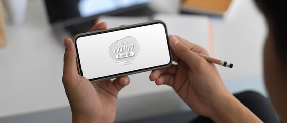 Hands holding mock up horizontal smartphone screen in blurred worktable background