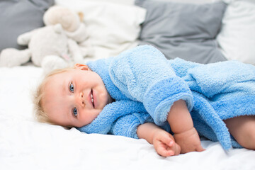 Ten month blond baby boy lying on bed in blue bathrobe and smiling. White pillows in background. Baby care concept, banner copy space