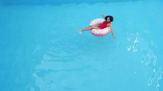 A young swimmer teenage girl sits in inflatable life-saving circle inblue clean cool water in sea pool, restores strength, makes waves on surface, moves her arms like butterfly rowing, distant plan