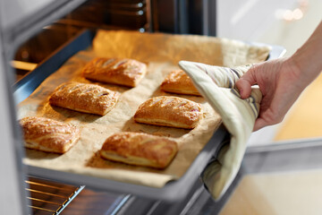 food cooking, culinary and people concept - young woman with potholder taking baking tray with jam pies out of oven at home kitchen