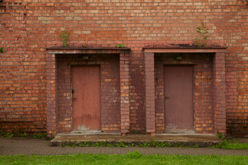 Wooden brown doors in an old red orange brick technical building, barn, warehouse. Grass and small trees grow on the roof.