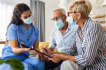 Female nurse talking to seniors patients with mask while being in a home visit, senior couple signs an insurance policy.