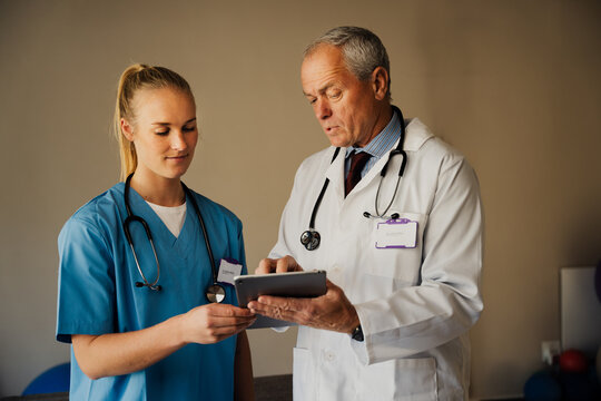 Male doctor teaching female intern in scrubs, while showing instructions on digital tablet in organised doctors room