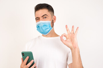Portrait of a happy Caucasian man wearing medical mask sending a message on his smartphone or taking a selfie isolated over white background and making ok sign with his hand.