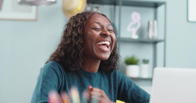 Beautiful female talking on video chat on computer while sitting indoor in room. Close up portrait of African American cheerful girl waving her hand and videochatting on laptop ay home Leisure concept