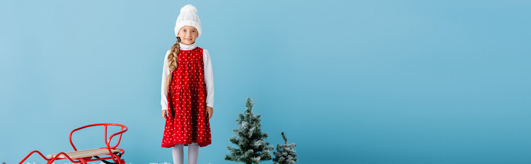 panoramic crop of girl in hat and winter outfit standing on near sleigh and pines isolated on blue