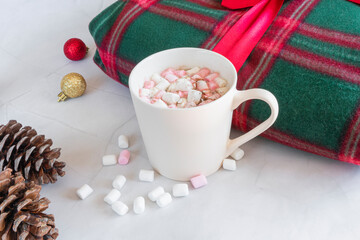 Obraz na płótnie Canvas Christmas hot drinks. White cup of hot coffee drink with marshmallows and cinnamon, warm plaid, pine cones, Christmas balls on a white marble background. Winter time. Christmas holidays concept