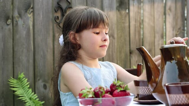 The girl is emotional and drinks tea from a cup. He takes a berry with his hand and eats it. Grimace, blinks and wipes his eyes with his hands. On the table is a plate of strawberries and a tea set.