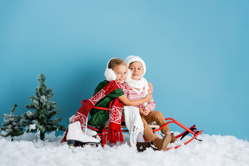 girl in winter earmuffs sitting on sleigh and hugging brother in hat near ice skates on blue