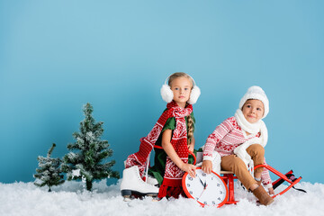 kids in scarfs and winter outfit sitting on sleigh near clock, pines and ice skates on blue