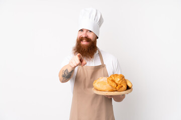 Redhead man in chef uniform. Male baker holding a table with several breads points finger at you with a confident expression
