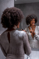 A young black girl with afro hair putting on makeup before the party with a smile. Exclusive party, vip party, gray evening dress