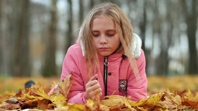 A little girl indifferently lies on the autumn foliage in a city park. She is very sad.