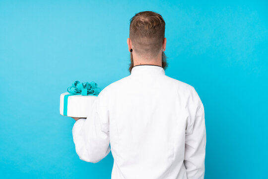 Redhead pastry chef with long beard holding a big cake over isolated blue background in back position