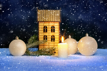 Christmas card with a lighted candle, Christmas balls and a wooden toy house during a snowfall