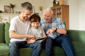 Smiling young handsome man resting on sofa with happy elderly father and small son, playing on...