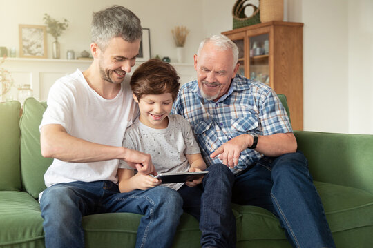 Smiling old grandpa, young grown son father and kid boy grandson watching or playing online at home on sofa