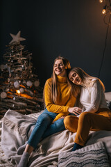 Obraz premium two girls in sweaters sit on the sofa in the New Year's room, one girl laid her head on the shoulder of the other.