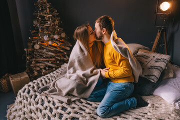 A romantic couple, sitting on the bed, kissing near the Christmas tree, wrapped in a blanket. Joyful moments of comfort during the winter holidays.