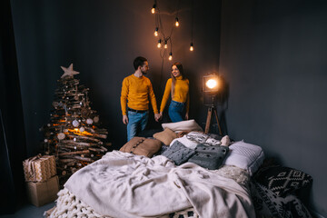 Loving couple with pillows on bed, garlands on the wall in a dark room with a warm yellow night light