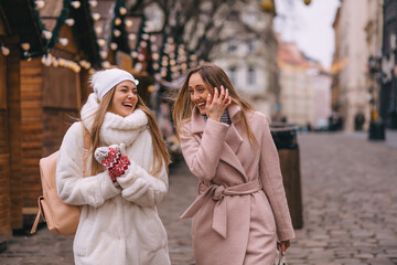 Fototapeta premium Two friends while shopping in knitted mittens, walking in the market square decorated with garlands on a winter day. Enjoy the winter festive mood.