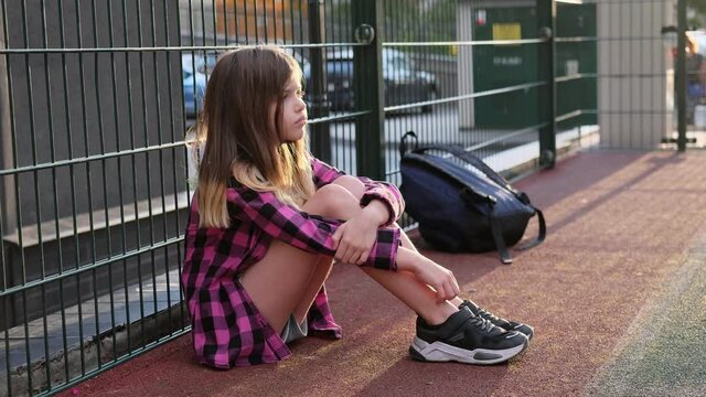 little / young lonely sad school girl in a pink plaid shirt with backpack sits alone on the street near the fence