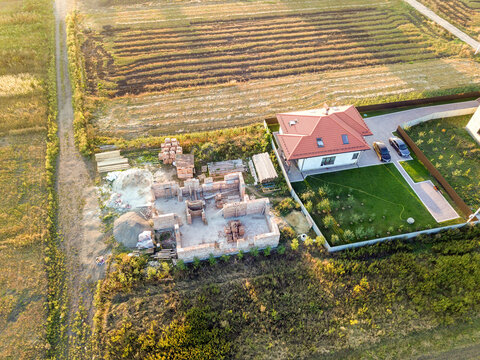 Top down aerial view of two private houses, one under construction with concrete foundament and brick walls and another finished with red tiled roof.
