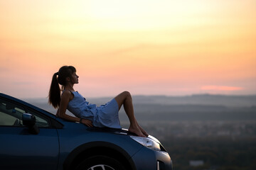 Happy young woman driver in blue dress enjoying warm summer evening laying on her car hood. Travelling and vacation concept.