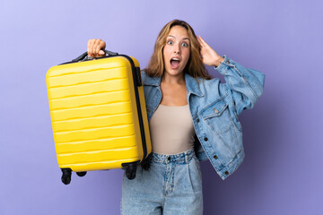 Young blonde woman isolated on purple background in vacation with travel suitcase and surprised