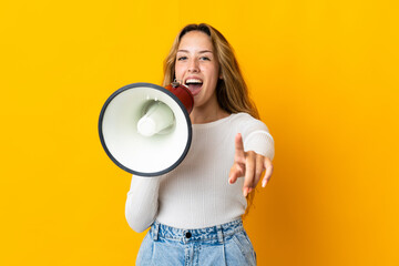Young blonde woman isolated on yellow background shouting through a megaphone to announce something while pointing to the front