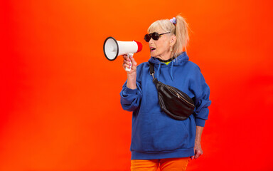 Shouting in megaphone. Senior woman in ultra trendy attire isolated on bright orange background. Looks stylish and fashionable, forever young. Caucasian model in sunglasses, bright attire and sneakers