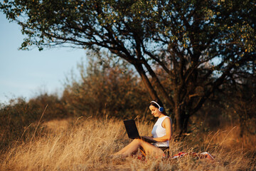 Naklejka na ściany i meble Young woman with laptop sitting on grass in park. She is texting on social networks on a laptop.