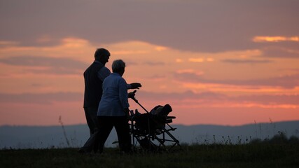 Couple of grandparents walk with baby carriage, beautiful sunset sky