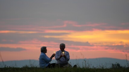 Fototapeta na wymiar Couple of old people holding sparkling fire work and singing, sunset sky, marriage anniversary