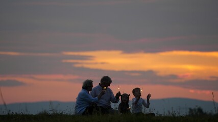 Fototapeta na wymiar Grandparent and children hold sparkling firework and singing, sunset sky