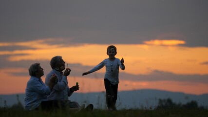 Fototapeta na wymiar Grandparents blowing soap bubbles while grandchildren play to catch them at sunset in the nature