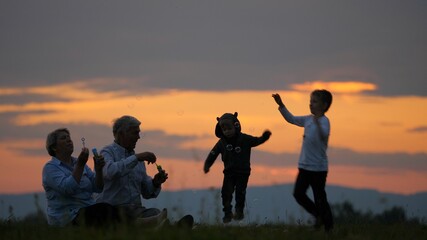 Fototapeta na wymiar Grandparent sit on hill and blow bubbles, children playing, sundown sky