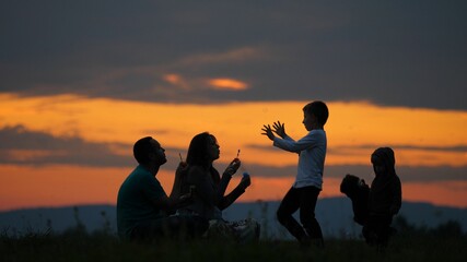 Fototapeta na wymiar Young parents silhouettes blowing bubbles and happy children play, red sunset sky in background