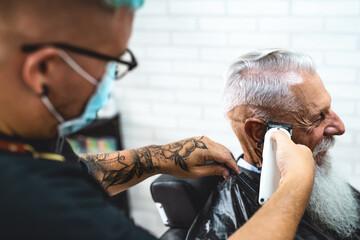 Male hairdresser cutting hair to hipster senior client while wearing face surgical mask - Young...