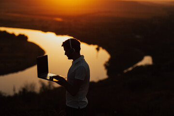 The silhouette of a man using a laptop on the background river and sunset. The guy combines rest with work.