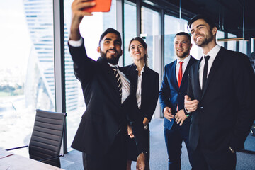 Successful cheerful male and female entrepreneurs taking selfie pictures on modern cellular phone during break of work in business center, smiling colleagues in formal wear posing while photographing