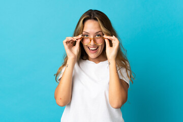 Young blonde woman isolated on blue background with glasses and surprised