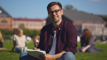 Obraz premium Portrait of handsome hipster guy student sitting on campus lawn with book looking at camera