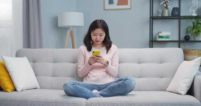 Girl sitting on couch and using smartphone in living room. Attractive young woman sending message, chatting with frien, family and laughing. Social media, online, Internet.